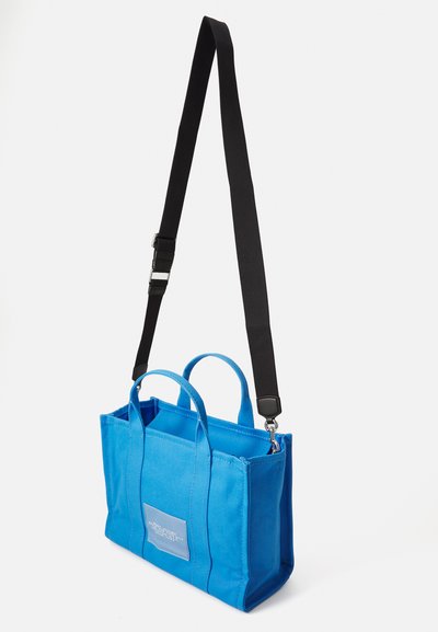 Sac en toile bleu avec deux poignées supérieures et une bandoulière noire réglable. Présente un patch sur le devant avec des détails textuels et une forme structurée.