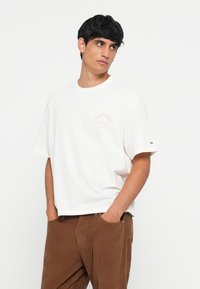 Camiseta blanca de algodón con cuello redondo, mangas cortas y un pequeño gráfico de color rosa claro en el pecho. Llevada con pantalones de pana marrón.