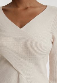 Helle beige gerippte Strickbluse mit V-Ausschnitt und Wickeldesign. Lange Ärmel und strukturiertes Gewebe erhöhen das visuelle Interesse und die Tiefe.