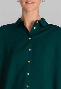 Camicia button-up verde scuro con una consistenza morbida, dotata di un colletto e sei bottoni metallici lungo la parte anteriore.