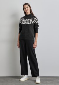 Maglione grigio scuro a maglia con motivo a zigzag bianco sul petto, con polsini a coste, abbinato a pantaloni larghi neri e sneakers bianche.