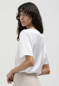 Blusa bianca a maniche corte con scollo rotondo, texture liscia, vestibilità morbida e leggero drappeggio sul retro, infilata in pantaloni beige.