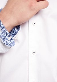 Weißes Hemd mit Knopfverschluss und Manschetten im blauen Blumenmuster. Die Manschette besteht aus einem glatten Stoff, während das Hemd schlicht und strukturiert ist.
