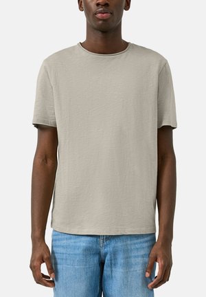 Homme portant un t-shirt uni gris clair à manches courtes et un jean bleu, debout devant un fond blanc uni.