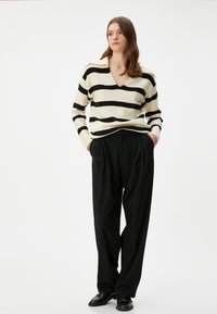 Gestreifter Pullover in Creme und Schwarz mit V-Ausschnitt und gerippten Manschetten, kombiniert mit lockeren schwarzen Hosen und schwarzen Schuhen.
