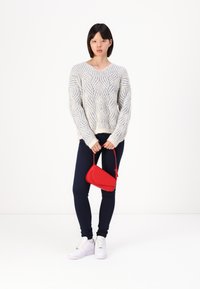 Lichtgrijze gebreide trui met golfpatroon, gecombineerd met marineblauwe legging en witte sneakers. Houdt een rode, driehoekige handtas vast.