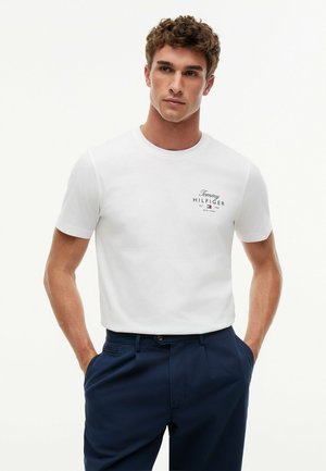 Junger Mann mit lockigem Haar, der ein weißes Tommy Hilfiger T-Shirt und dunkelblaue Hose trägt, steht mit den Händen in den Taschen vor einem schlichten Hintergrund.