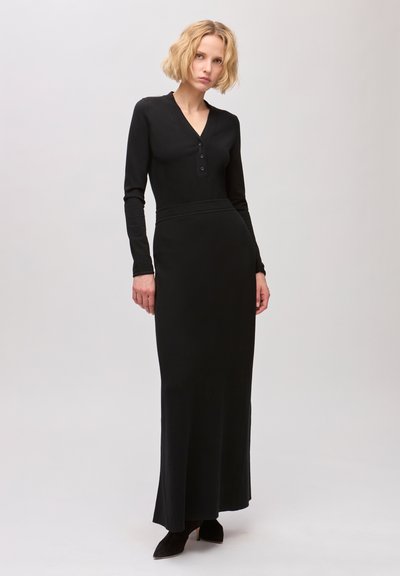 Femme aux cheveux blonds courts portant une longue robe noire ajustée avec manches longues et encolure boutonnée, debout devant un fond clair uni.