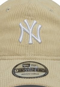 Căciulă bej din corduroy cu logo-ul alb brodat "NY". Dispune de o curea ajustabilă și o etichetă circulară cu logo-ul care indică "New Era 9TWENTY."