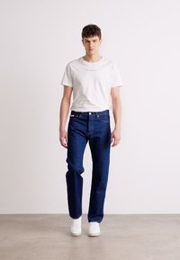 Camiseta blanca con texto, combinada con jeans de mezclilla azul oscuro. El look se completa con unas zapatillas blancas. Diseño simple y casual.