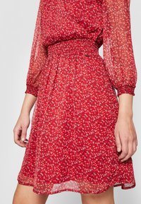 Vestido rojo con pequeño estampado floral blanco, cintura elástica y mangas largas translúcidas con puños fruncidos, sobre una persona de pie.