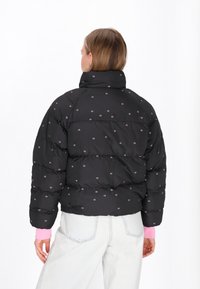 Sort pufferjakke med lyserøde blomster mønstre, høj krave og quiltet design. Båret over en lyserød langærmet skjorte og blå jeans.