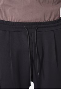 Pantaloni neri con vita regolabile, pieghe e una texture liscia. La parte superiore mostrata è una camicia corta di colore marrone tenue.