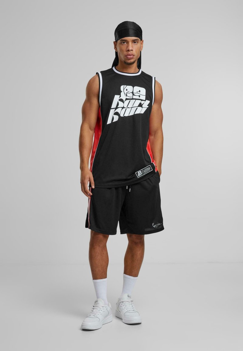 Bărbat care poartă un tricou sport negru fără mâneci cu detalii albe și roșii, pantaloni scurți negri, șosete albe, adidași albi și un batic negru pe cap, stând pe un fundal gri.