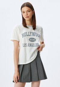 Camiseta gráfica blanca con la inscripción "HOLLYWOOD EST 1991 LOS ÁNGELES" en gris, combinada con una falda plisada gris. Corte casual, mangas cortas.