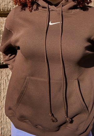 Person wearing a brown Nike hoodie with drawstrings and a front pocket, standing against a rough stone wall.