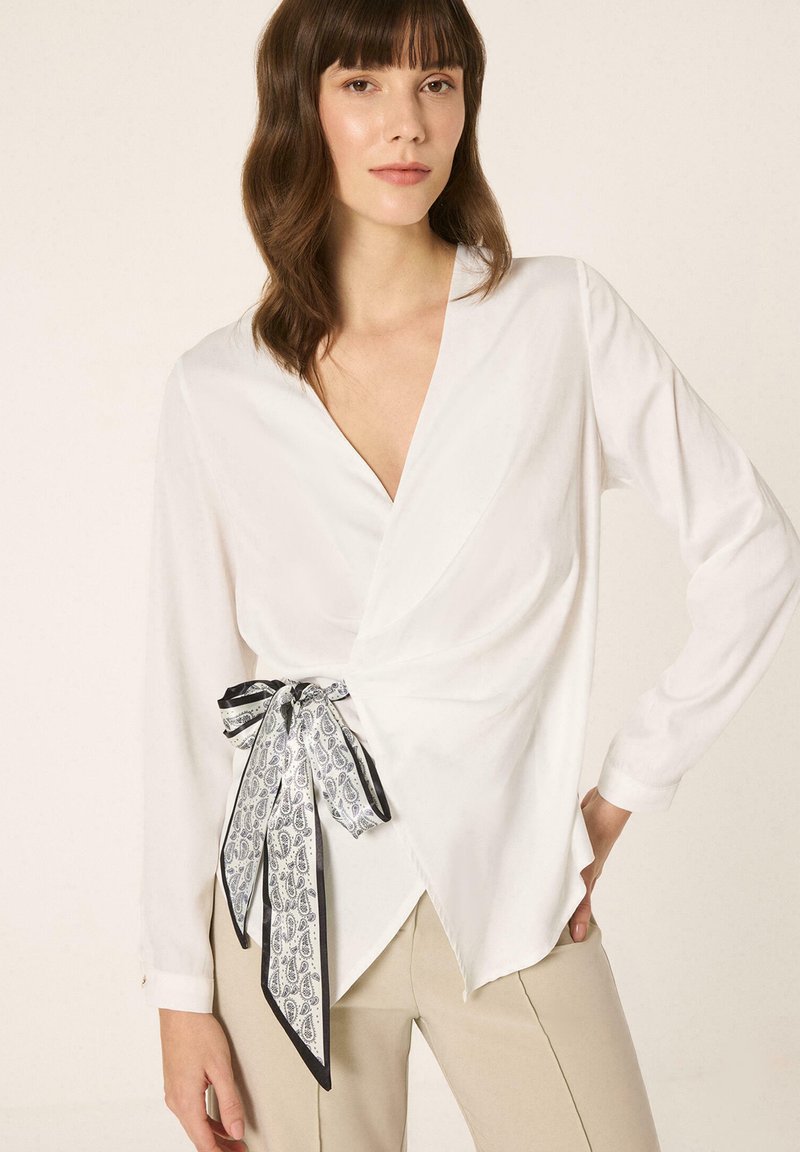 Femme portant un chemisier cache-cœur blanc avec un nœud à motif cachemire noir et blanc, associé à un pantalon beige, debout avec une main sur la hanche.