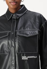 Giacca camicia in ecopelle nera con cuciture bianche, chiusura a bottoni, due tasche sul petto e "KARL LAGERFELD JEANS" ricamato sul davanti.