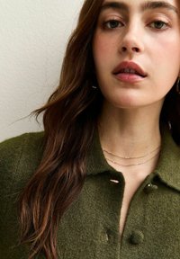 Joven con cabello largo y castaño, vistiendo una camisa verde con textura y botones, y un delicado collar de cadena dorada, sobre un fondo claro y liso.