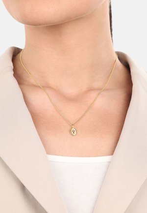 Goldene Halskette mit einem ovalen Anhänger, der ein Kreuzdesign zeigt, aufgehängt an einer zarten Kette, getragen über einem hellen Oberteil.