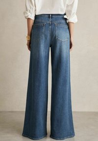 Jeans in denim a gamba larga in un lavaggio blu scuro, caratterizzati da un alto punto vita, tasche posteriori e dettagli di cuciture sottili. Tessuto dalla texture liscia.