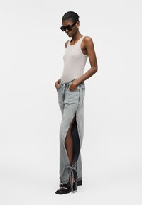 Mujer con top sin mangas beige, vaqueros grises de pierna ancha con abertura hasta el muslo atados en los tobillos, zapatos negros de tacón y gafas de sol oscuras, de pie con las manos en los bolsillos.