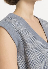 Hedvábný bezrukávový top s modrým vzorem, který má kostkovaný design, texturovanou tkaninu a hluboký V-výstřih, zobrazující různé odstíny modré.