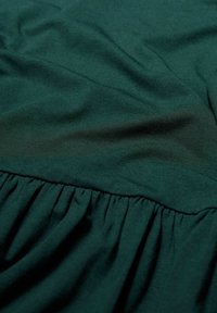 Ciemnozielony materiał o gładkiej fakturze, z marszczonymi falbanami wzdłuż dolnej krawędzi i płaską, kontrastową wstawką na górze.