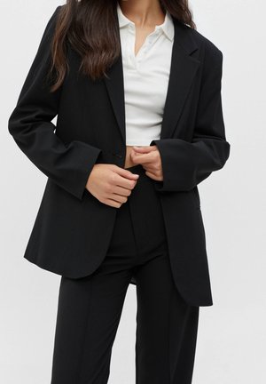 Femme portant un blazer noir et un pantalon avec un polo court blanc, mains ajustant le blazer au niveau de la taille.