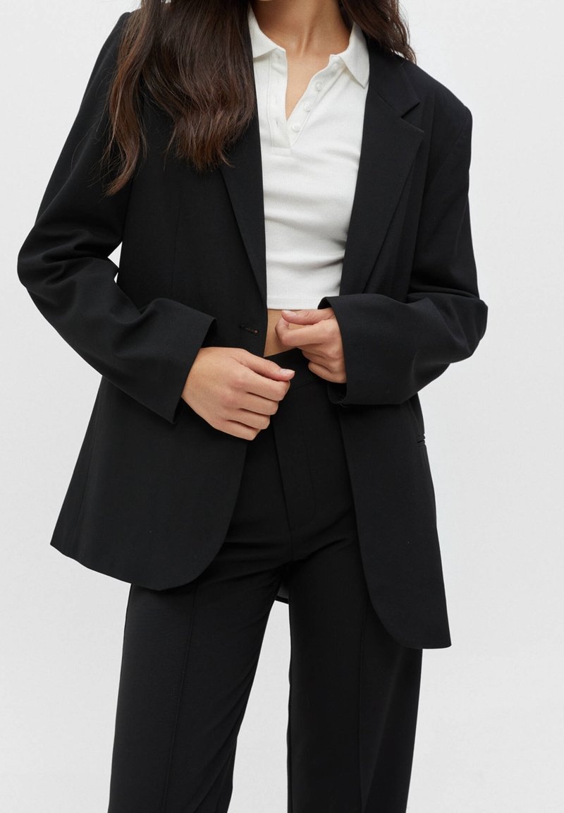 Femme portant un blazer noir et un pantalon avec un polo court blanc, mains ajustant le blazer au niveau de la taille.