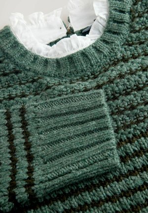 Zelený svetr s vzorem pletený přes bílou blůzu s volány, přičemž jsou viditelné podrobné textury a žebrované manžety.