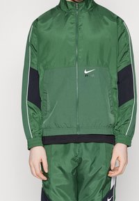 Giacca a zip Nike verde con pannello centrale testurizzato, accenti neri sulle maniche e piping bianco lungo i bordi. Chiusura frontale a zip.