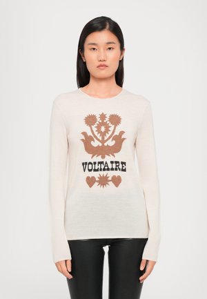 Pull crème à manches longues avec un design graphique brun orné de motifs floraux et du mot "VOLTAIRE" en noir, associé à un pantalon noir.
