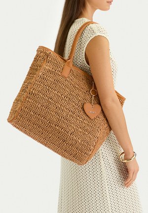 Mujer vestida con un vestido blanco de crochet que sostiene un gran bolso tote marrón tejido con asas de cuero y un charm en forma de corazón.