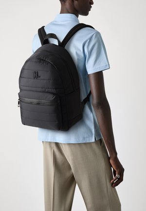 Mochila acolchada negra con forma redondeada, bolsillo frontal con cremallera y tirantes ajustables, llevada sobre una camisa azul clara y pantalones beiges.