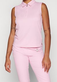 Ljust rosa ärmlös polo skjorta med krage, framdragkedjedetalj och texturerad tyg. Matchas med ljust rosa figurnära byxor.