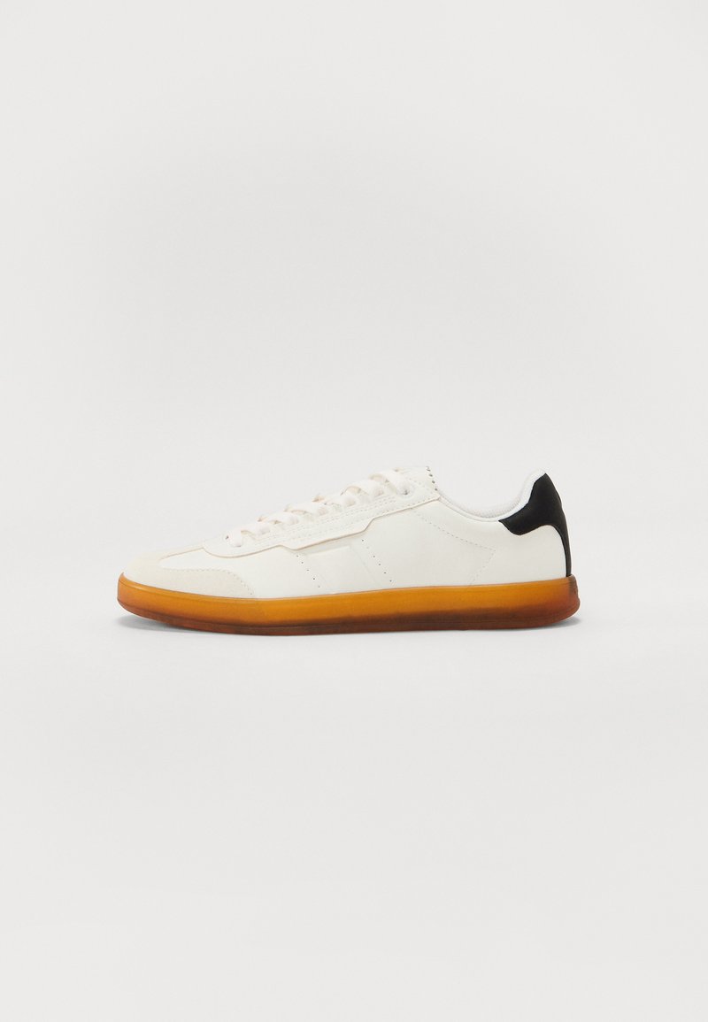 White sneaker with a smooth leather upper, black heel accent, and orange rubber sole; features laces and minimalistic design lines.
