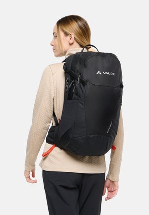 Femme portant une veste beige et un pantalon noir, portant un grand sac à dos noir Vaude avec plusieurs compartiments et des sangles rouges.