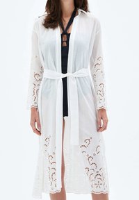 Biała, lekka narzutka z długim rękawem, z wiązaniem w talii. Posiada misternie haftowaną koronkę na rękawach i u dołu.