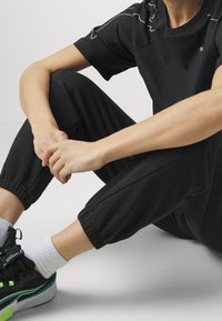 Persoon zittend op de vloer, gekleed in zwarte joggers, een zwart T-shirt met opdruk, witte sokken en zwarte sneakers met neon groene accenten.