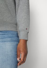 Sudadera gris con puños acanalados, que muestra un pequeño parche de logo en rojo, blanco y azul en la manga. Combinada con jeans azules.