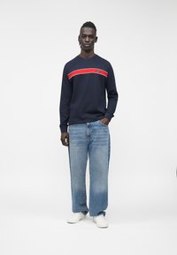 Felpa blu navy a coste con una striscia orizzontale rossa e bianca, abbinata a jeans a gamba larga color azzurro chiaro e sneakers bianche. Sfondo bianco semplice.