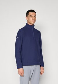 Pullover azul marinho com meia-zíper feito de tecido texturizado, com mangas longas e um logótipo na manga esquerda. Corte casual e cor sólida.