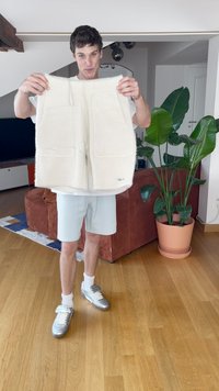Kremasto obarvan, puhast tkaninski jopič z dvema sprednjima žepoma, ki ga drži oseba v svetlih kratkih hlačah. Notranji prostor z zelenimi rastlinami in lesenim podom.