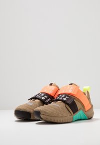 Sneaker mit einem oberen Teil aus braunem Mesh, schwarzen und orangefarbenen Riemen, grünen Akzenten auf der Sohle und einer strukturierten Oberfläche. Leichtes Design.