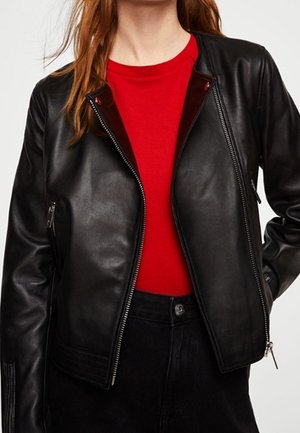 Frau trägt eine offene schwarze Lederjacke über einem eng anliegenden roten Shirt und schwarzen Hosen vor einem schlichten Hintergrund.