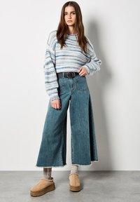 Niepowtarzalny, niebiesko-biały sweter w paski, spodnie dżinsowe z wysokim stanem i szerokimi nogawkami z frędzlami, oraz beżowe półbuty wsuwane na grubym spodzie.