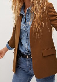 Blazer marron au design ajusté, associé à une chemise en denim bleu clair. Tissu doux, poche avant simple et ceinture noire.