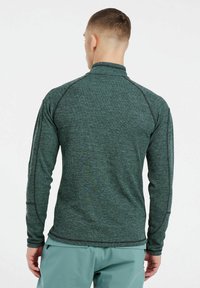 Grünes Fleece-Oberteil mit langen Ärmeln und hohem Kragen, das über kontrastierende Nähte und einen strukturierten Stoff verfügt. Die Rückansicht zeigt eine enganliegende Passform und einen athletischen Schnitt.