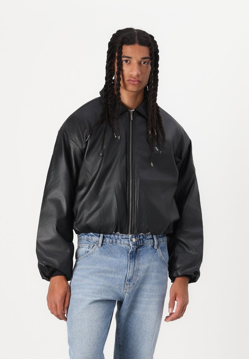 Giubbotto bomber in ecopelle nera con chiusura a zip, vestibilità oversize, polsini elasticizzati e colletto rotondo, abbinato a jeans chiari.